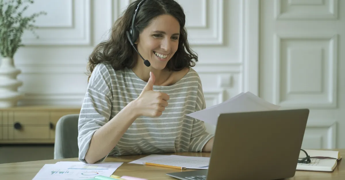 Top Remote Jobs for 2025 - A professional woman working remotely from home, wearing a headset, giving a thumbs-up while looking at her laptop.