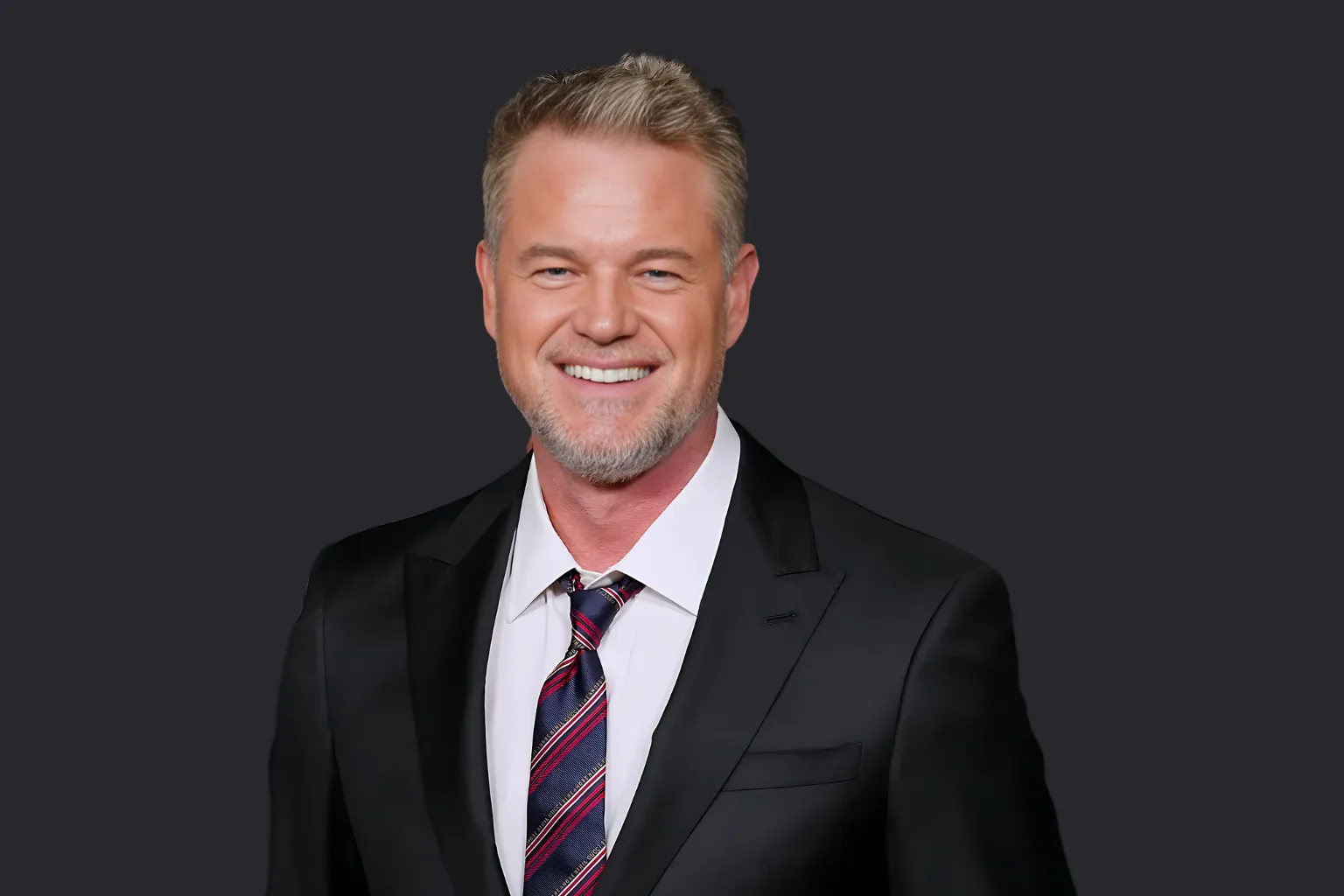 Eric Dane smiling in a black suit with striped tie, representing his USA entertainment legacy of inspiring triumphs and tough trials.
