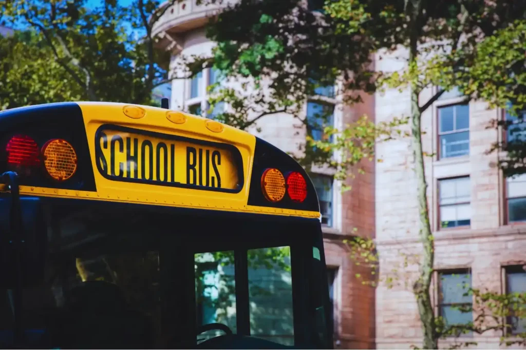 School Bus Awaiting Students for USA School Day The rear view of a yellow American school bus, with its "SCHOOL BUS" sign illuminated, parked in front of an old, classic school building with green trees in the foreground. This image symbolizes the routine of US school days.