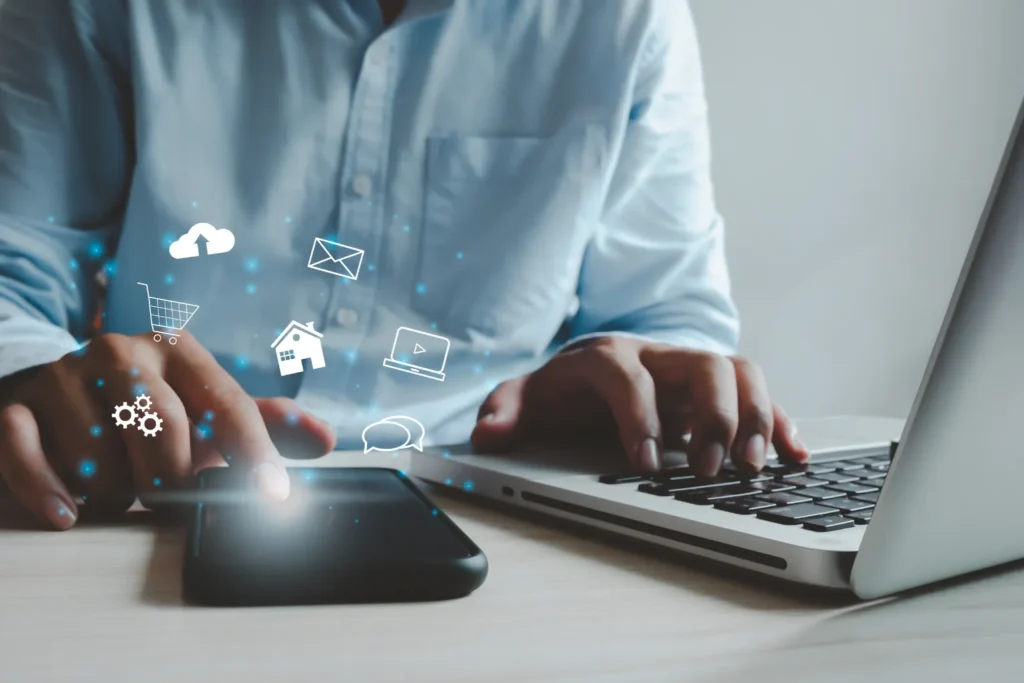 Close-up of hands typing on a laptop with a smartphone nearby, surrounded by glowing digital icons for cloud, email, shopping, and media, symbolizing work-from-home flexibility.