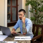 Man working on laptop and phone while how to start a small online business from home with documents and workspace setup