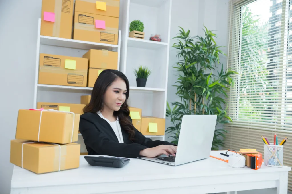 Start a Small Online Business from Home with eCommerce Setup Woman managing online business from home packing orders and working on laptop with boxes and shipping supplies